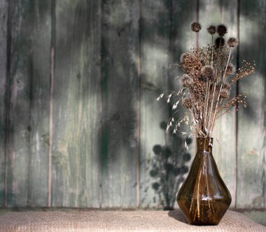 Ahşap duvarın arka planında bir buket kuru çiçek ve cam vazoda bitkiler. Kırsal tarzda sakin bir hayat. Sert ışık. Metin için yeri olan kartpostal.