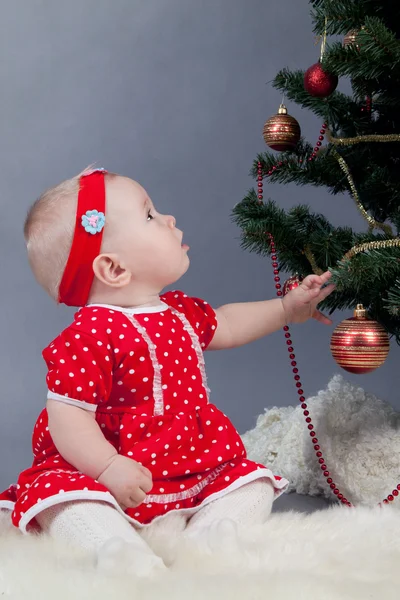 Klein meisje in een rode jurk zitten in de buurt van de kerstboom — Stockfoto