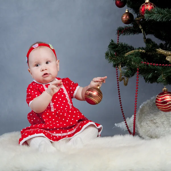 Kleines Mädchen in rotem Kleid sitzt neben Weihnachtsbaum — Stockfoto