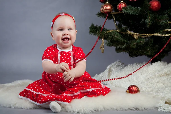 Klein meisje in een rode jurk zitten in de buurt van de kerstboom — Stockfoto