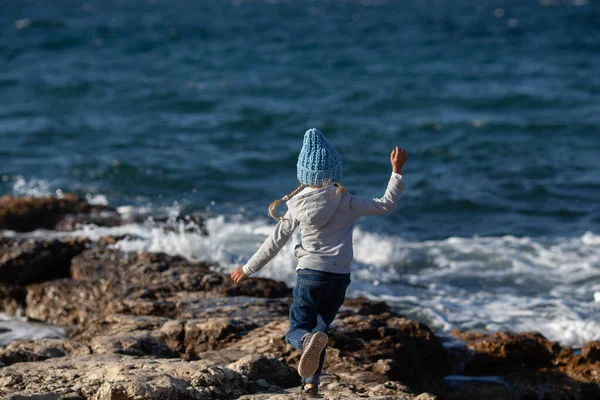 在大海的背景下 在美丽的风景后面 一个快乐漂亮的姑娘沿着海滨散步 那孩子戴着一顶蓝色针织的帽子 — 图库照片