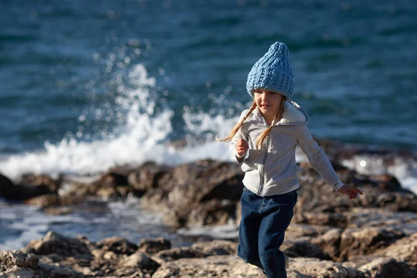 美しい風景の後ろから 海の背後にある海の海岸に沿って幸せなかなりの女の子歩く 子供は青いニットの帽子をかぶっている — ストック写真