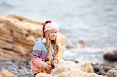 Deniz kıyısında Noel Baba şapkalı küçük güzel kız, ceketli bir çocuk yürüyor.