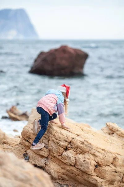 海岸のサンタ クラスの帽子の可愛い女の子子供はジャケットの中を歩く — ストック写真