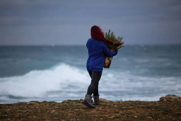 一个红头发的漂亮女孩拿着一个装有圣诞树树枝的手袋 一个女人穿着一件蓝色的毛衣 沿着冬季的海滨散步 — 图库照片