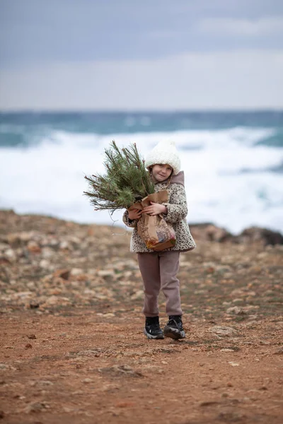 一个漂亮的女孩拿着一个装有圣诞树树枝的手袋 这孩子穿着毛皮外套 头戴白帽 沿着冬季的海滨散步 — 图库照片