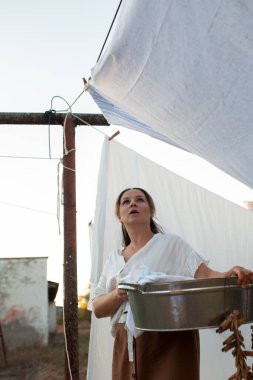 Uzun saçlı güzel bir kadın deniz fenerinin yanındaki havaya beyaz çarşaflar asıyor. Sıcak bir sonbahar günü, günbatımı, akşam, açık hava.