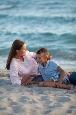 Anne ve oğlu mavi deniz manzarasında birbirlerine sarılıp gülümsüyorlar.