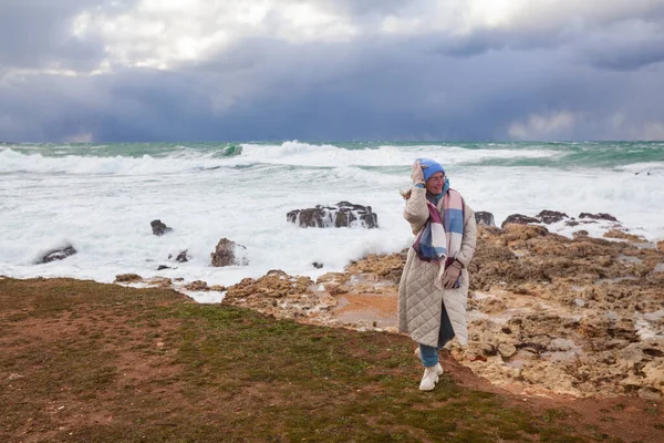 一个时髦的女人走在冬季的海面上 她穿着米黄色的外套 戴着一顶帽子 海上刮起了风暴 — 图库照片