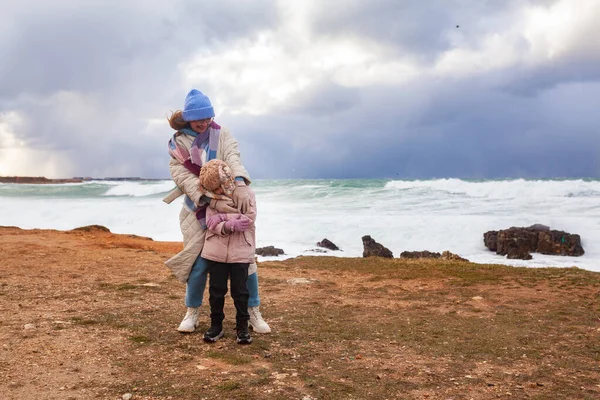 快乐的妈妈和女儿在寒风凛凛的海面上散步 一个女人和一个女孩抱在怀里笑着 — 图库照片