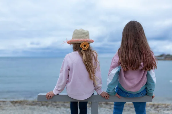 一个朋友的两个女孩坐在一个金属结构上 看着大海 孩子们穿着牛仔裤和一件连衣裙 — 图库照片