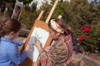 Kız parkta bir sehpaya resim çiziyor, plein havası. Çocuk öğretmenle birlikte doğada resim yapmayı öğrenir. Öğretmen açıklar ve nasıl çizileceğini gösterir.