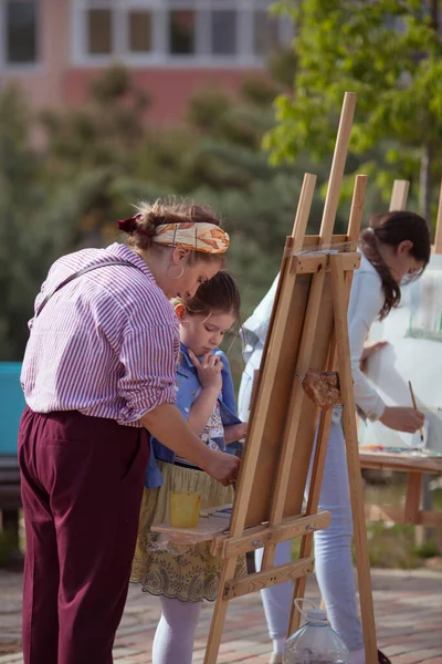 Kız parkta bir sehpaya resim çiziyor, plein havası. Çocuk öğretmenle birlikte doğada resim yapmayı öğrenir. Öğretmen açıklar ve nasıl çizileceğini gösterir.