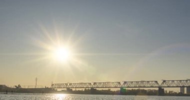 Şehir nehir kıyısının zaman çizelgesi. Güneş ışınları, mavi gökyüzü ve ufukta demiryolu köprüsü. Düz küçük dalgalar, su ve motorlu kaydırak hareketleriyle yaz günbatımı zamanı