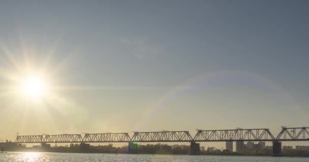 Timelapse brzegu rzeki miasta. Promienie słoneczne, błękitne niebo i most kolejowy nad horyzontem. Letni zachód słońca z płaskimi, malutkimi falami wody i zmotoryzowanym ruchem suwaka — Wideo stockowe