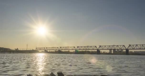 Тімелапс міського берега річки. Сонячні промені, блакитне небо і залізничний міст над горизонтом. Літній час заходу сонця з пласкою крихітною хвилею води та рухом моторизованого повзунка — стокове відео