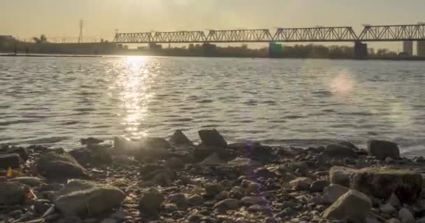 A városi folyópart időrendje. Napsugarak, kék ég és vasúti híd a horizont felett. Nyári naplementekor lapos apróhullámok víz és motoros csúszó mozgás — Stock videók