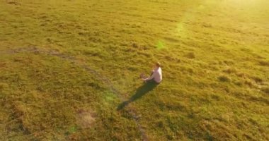 Havadan bakış açısı. Yeşil çimlerde oturan ve kırsal alanda not defteri olan bir adamın etrafında alçak yörünge uçuşları. Günlük giysi ve zen meditasyon pozu. Güneş ışınları. Radikal hareket.