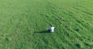 Havadan bakış açısı. Yeşil çimlerde oturan ve kırsal alanda not defteri olan bir adamın etrafında alçak yörünge uçuşları. Günlük giysi ve zen meditasyon pozu. Güneş ışınları. Radikal hareket.