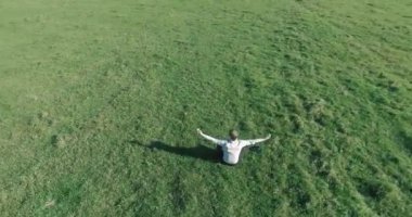 Havadan bakış açısı. Yeşil çimlerde oturan ve kırsal alanda not defteri olan bir adamın etrafında alçak yörünge uçuşları. Gündelik kıyafet ve Zen meditasyonu. Eller havada. Güneş ışınları. Radikal hareket.