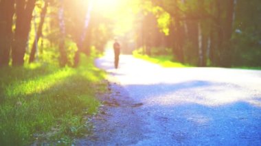Yeni asfalt yolda koşan FHD sporcusu. Şehir parkında gün doğumu ya da gün batımı. Yeşil ağaç ormanı ve ufukta güneş ışığı.