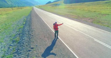 Hava 4K UHD görüntüsü. Asfalt yolda yürüyen otostopçu turist üzerinde alçak irtifa uçuşu. Büyük bir kırsal vadi ve yaz günü güneşli bir çayır. Sırt çantalı gezgin..
