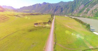 Hava UHD 4K: güneşli yaz sabahında kırsal dağ yolu ve çayır üzerinde havadan uçuş. Ufukta yeşil ağaçlar ve güneş ışınları var. Özgür kırsal asfalt karayolu ve nehir.