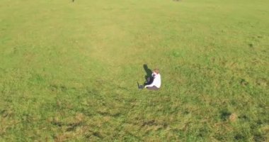 Havadan bakış açısı. Yeşil çimlerin üzerinde oturan, sarı kırsal alanda not defteri olan ve telefonla konuşan bir adam. Günlük kıyafet. Güneş ışınları. Radikal hareket.