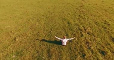 Havadan bakış açısı. Yeşil çimlerde oturan ve kırsal alanda not defteri olan bir adamın etrafında alçak yörünge uçuşları. Gündelik kıyafet ve Zen meditasyonu. Eller havada. Güneş ışınları. Radikal hareket.