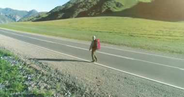 Hava 4K UHD görüntüsü. Asfalt yolda yürüyen otostopçu turist üzerinde alçak irtifa uçuşu. Büyük bir kırsal vadi ve yaz günü güneşli bir çayır. Sırt çantalı gezgin..