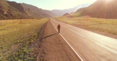Hava 4K UHD görüntüsü. Asfalt yolda yürüyen otostopçu turist üzerinde alçak irtifa uçuşu. Büyük bir kırsal vadi ve yaz günü güneşli bir çayır. Sırt çantalı gezgin..