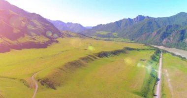 Hava UHD 4K: güneşli yaz sabahında kırsal dağ yolu ve çayır üzerinde havadan uçuş. Ufukta yeşil ağaçlar ve güneş ışınları var. Özgür kırsal asfalt karayolu ve nehir.