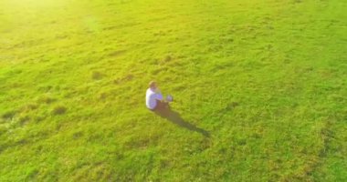 Havadan bakış açısı. Yeşil çimlerin üzerinde oturan, sarı kırsal alanda not defteri olan ve telefonla konuşan bir adam. Günlük kıyafet. Güneş ışınları. Radikal hareket.