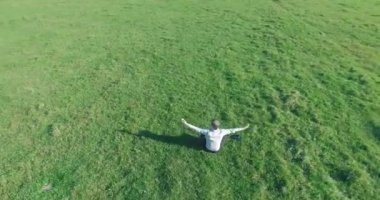 Havadan bakış açısı. Yeşil çimlerde oturan ve kırsal alanda not defteri olan bir adamın etrafında alçak yörünge uçuşları. Gündelik kıyafet ve Zen meditasyonu. Eller havada. Güneş ışınları. Radikal hareket.