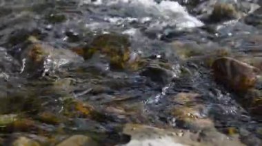 Ormanın yakınındaki küçük bir dağ nehrinde akan suyun motorlu bebek kaydırağıyla çekilmiş görüntüsü. Kocaman ıslak kayalar ve güneş ışınları. Kırsal bölgede yatay sabit hareket. Çiğ düz renkler.