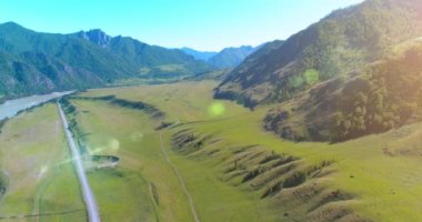 Hava UHD 4K: güneşli yaz sabahında kırsal dağ yolu ve çayır üzerinde havadan uçuş. Ufukta yeşil ağaçlar ve güneş ışınları var. Özgür kırsal asfalt karayolu ve nehir.