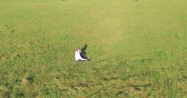 Havadan bakış açısı. Yeşil çimlerin üzerinde oturan, sarı kırsal alanda not defteri olan ve telefonla konuşan bir adam. Günlük kıyafet. Güneş ışınları. Radikal hareket.