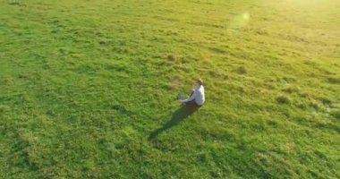 Havadan bakış açısı. Yeşil çimlerde oturan ve kırsal alanda not defteri olan bir adamın etrafında alçak yörünge uçuşları. Günlük giysi ve zen meditasyon pozu. Güneş ışınları. Radikal hareket.