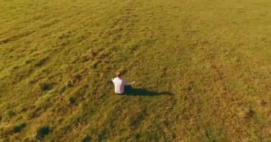 Havadan bakış açısı. Yeşil çimlerde oturan ve kırsal alanda not defteri olan bir adamın etrafında alçak yörünge uçuşları. Günlük giysi ve zen meditasyon pozu. Güneş ışınları. Radikal hareket.