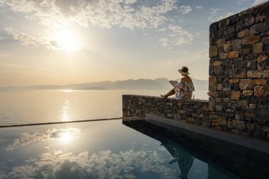 Rahatlama ve meditasyon kavramı - kadın lüks bir otelde gün doğumunda deniz manzaralı bir havuz kenarında kitap okuyor.