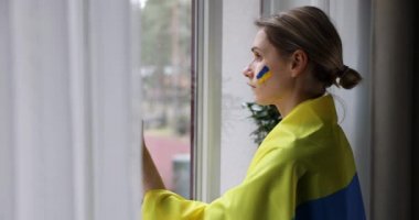 Yanağında ve omuzlarında Ukrayna bayrağı olan üzgün kadın pencereden dışarı bakıyor. destek, dayanışma ve empati kavramı