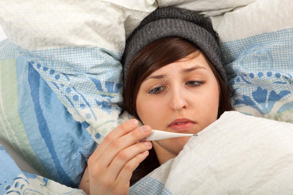 The young girl is lying sick in bed and taking her temperature