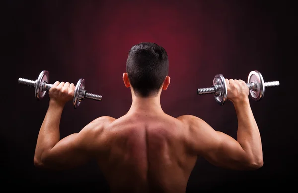 Musculoso joven levantando pesas desde la espalda contra el respaldo negro — Foto de Stock