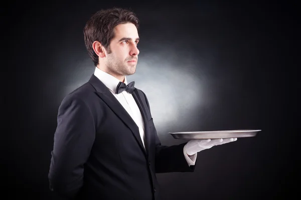 Portrait of a young waiter holding an empty dish on black backgr — Stock Photo, Image