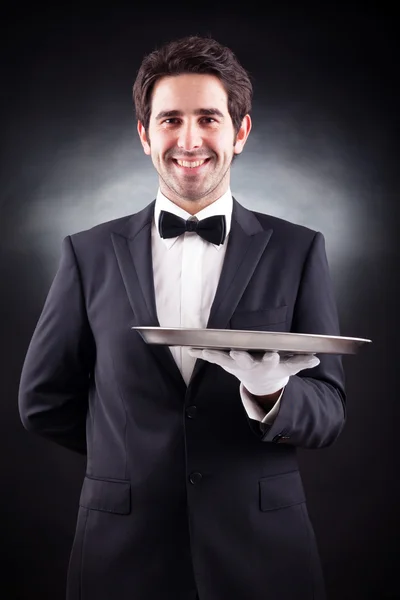 Portrait of a young waiter holding an empty dish on black backgr — Stock Photo, Image