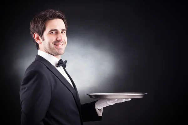 Portrait of a young waiter holding an empty dish on black backgr — Stock Photo, Image