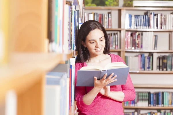 Porträt einer lächelnden jungen Studentin, die in einer Bibliothek ein Buch liest — Stockfoto