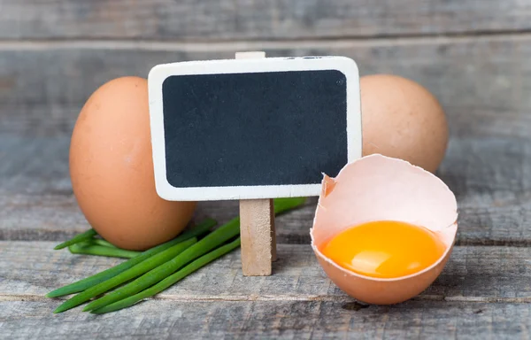 Whole and broken brown eggs with fresh onion and small blackboard — Stock Photo, Image