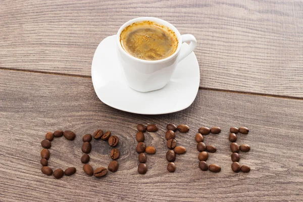Šálek kávy a slovo kávy z kávových zrn dřevěný stůl — Stock fotografie