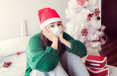 Noel için kilitlenme. Yüzü maskeli ve Noel ağacının arkasında Noel Baba şapkası takan kız.